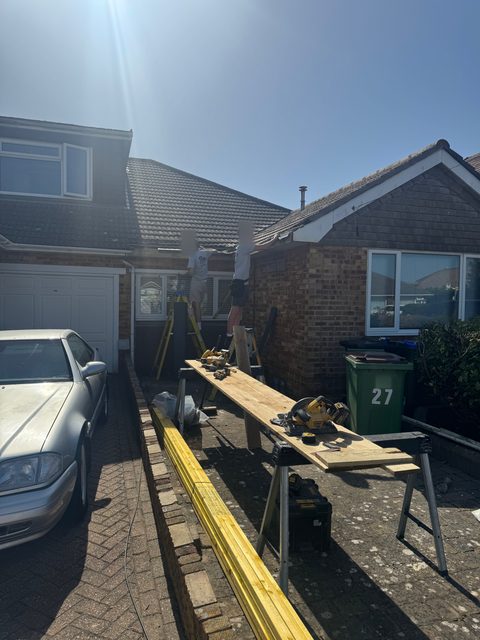 Storm Dry Roofing team working at the eaves of a bungalow, with new timber being cut on trestles on the driveway
